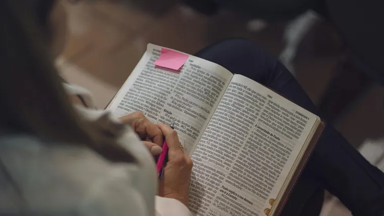 Uma mulher lendo uma Bíblia com um bilhete rosa em um ambiente interno, em devoção e estudo.