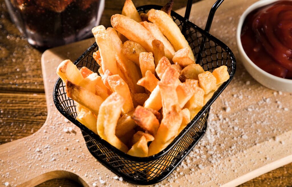 Cooked French fries in a black wire food basket with ketchup nearby