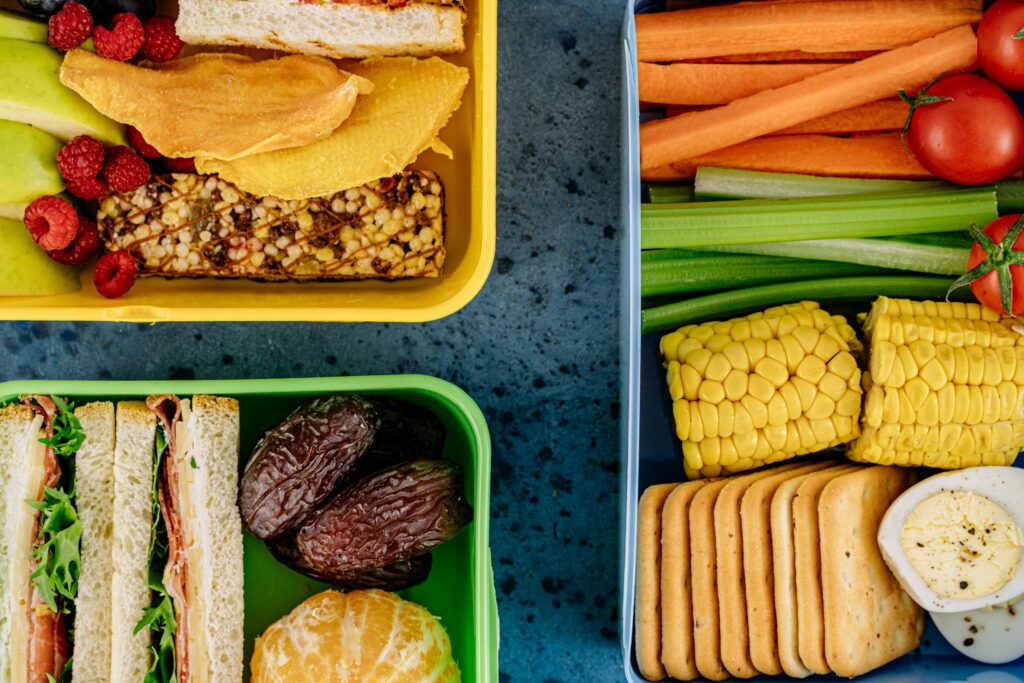 Lunch containers filled with sandwiches, fruit, carrots, celery, corn, crackers, and egg