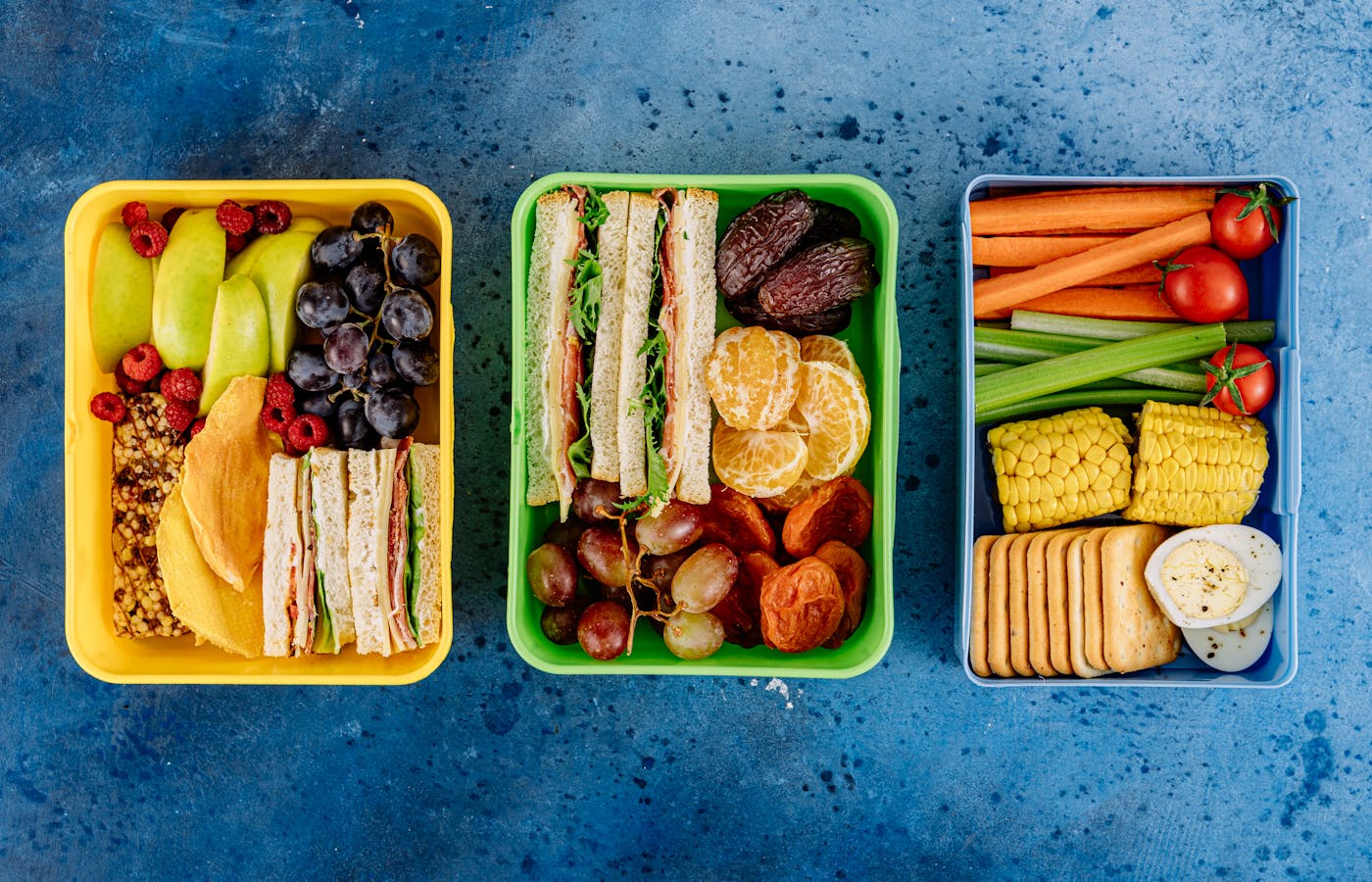 Three colorful lunch boxes filled with sandwiches, fruit, vegetables, crackers, and eggs