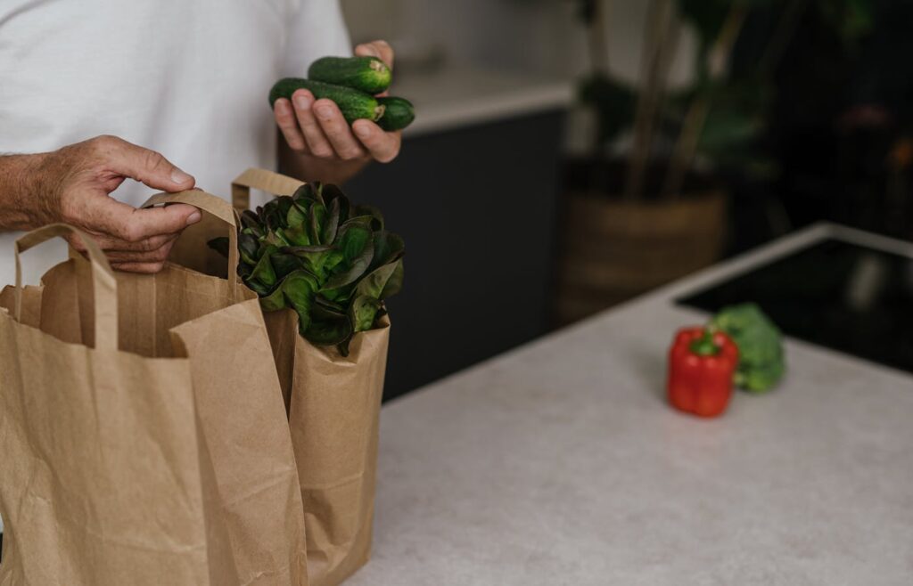 Hands unpacking cucumbers and artichokes from paper grocery bags on a kitchen counter